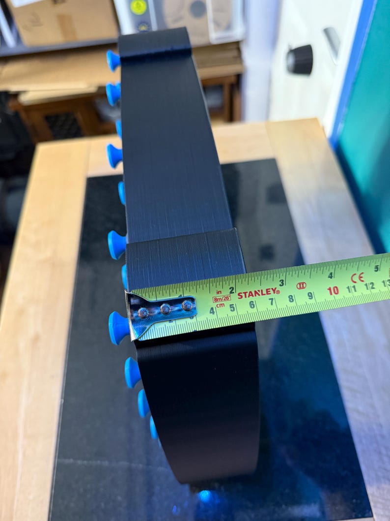 May include: A black, curved object with blue knobs along the edge. A yellow and green tape measure shows a width of approximately 2 inches. The object appears to be made of metal.