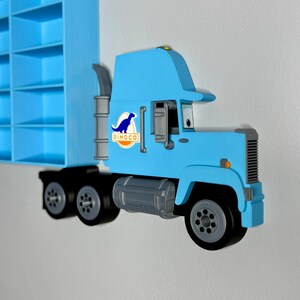 May include: A light blue toy truck with black wheels and a gray exhaust pipe. The truck features the Dinoco logo on the side. A blue shelf is visible in the background.