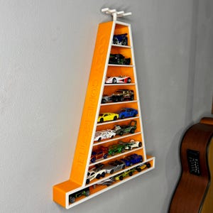 May include: A wall-mounted shelf in the shape of a cone, coloured orange and white, displaying toy cars. The words "COZY CONE MOTEL" are printed on the side. The shelf is fixed to a grey wall.