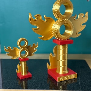 May include: Two gold-coloured trophies with a tyre and flame design. The trophies have a red base and a gold column. The larger trophy has the word "WINNER" on the base. The background is a teal colour.