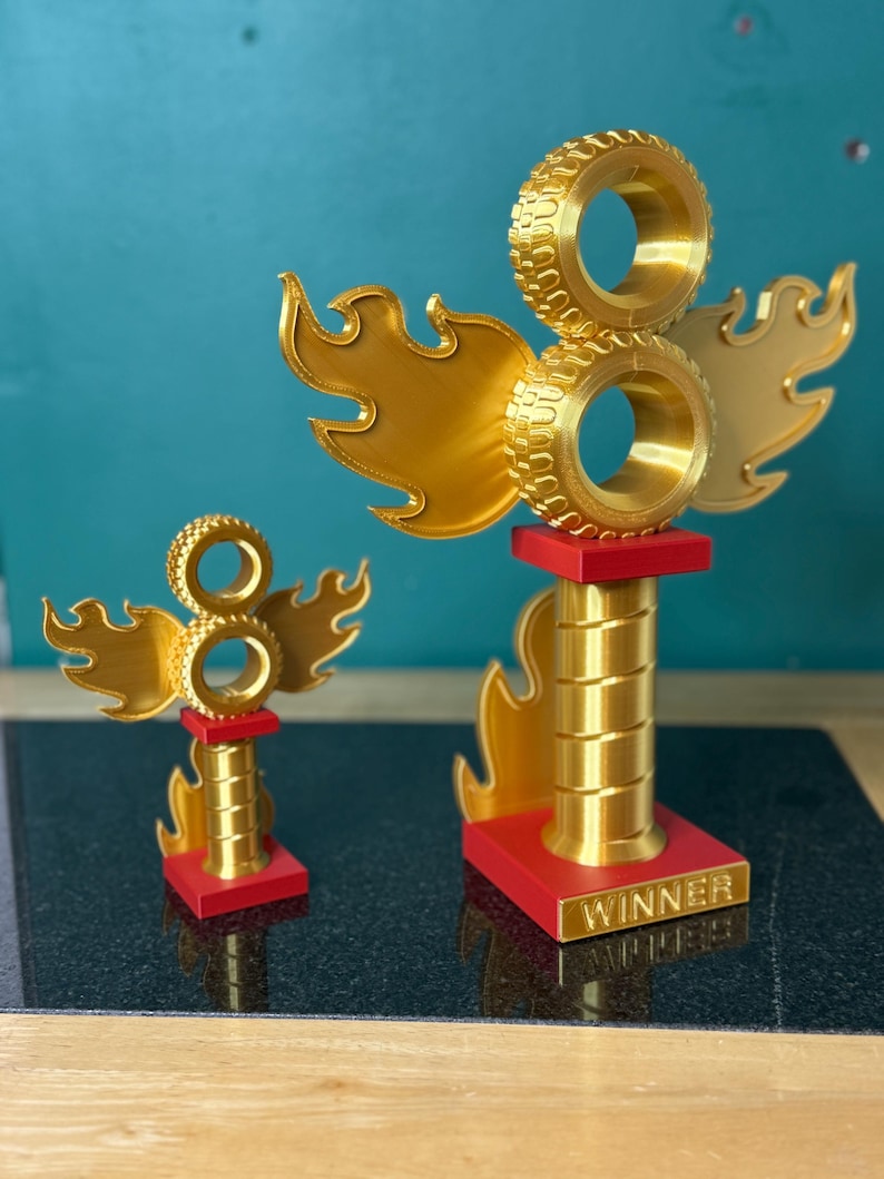 May include: Two gold-coloured trophies with a tyre and flame design. The trophies have a red base and a gold column. The larger trophy has the word "WINNER" on the base. The background is a teal colour.