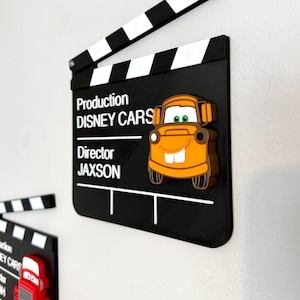 May include: Black film clapperboard with the text "Production DISNEY CARS Director JAXSON" and a 3D orange cartoon car. The clapperboard has a white and black top. Another clapperboard with a red cartoon car is visible.