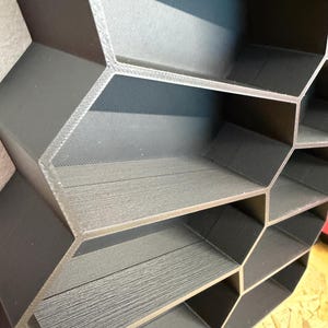 May include: A dark gray honeycomb-shaped wall shelf unit featuring multiple hexagonal compartments. The structure is constructed with a textured, layered finish, creating a geometric grid pattern with open storage spaces for organizing small items.