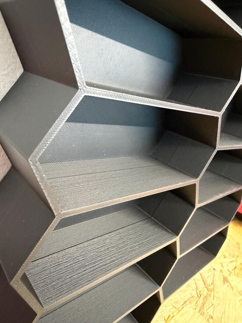 May include: A dark gray honeycomb-shaped wall shelf unit featuring multiple hexagonal compartments. The structure is constructed with a textured, layered finish, creating a geometric grid pattern with open storage spaces for organizing small items.