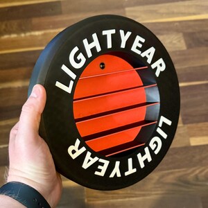 May include: Black and red circular decorative item with the word "LIGHTYEAR" in white lettering. The item has a series of horizontal red lines in the center, and is held by a hand.