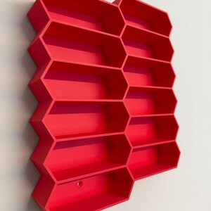 May include: A bright red wall-mounted storage unit featuring a honeycomb-inspired design with multiple hexagonal compartments. The structure is composed of interconnected geometric cells arranged in a vertical pattern, providing open shelving space.