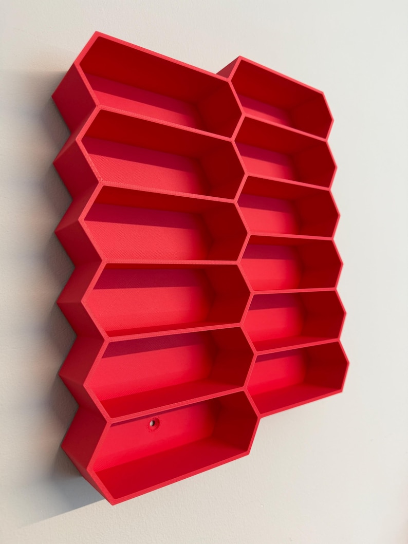 May include: A bright red wall-mounted storage unit featuring a honeycomb-inspired design with multiple hexagonal compartments. The structure is composed of interconnected geometric cells arranged in a vertical pattern, providing open shelving space.