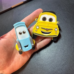 May include: Two cartoon car-shaped magnets. One is yellow with a black top and black tires, the other is light blue with yellow accents and silver details. Both have smiling faces and large eyes. The magnets are held in a hand.