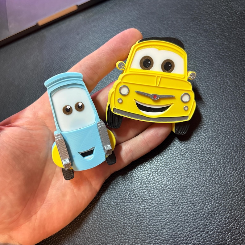 May include: Two cartoon car-shaped magnets. One is yellow with a black top and black tires, the other is light blue with yellow accents and silver details. Both have smiling faces and large eyes. The magnets are held in a hand.