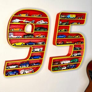 May include: Two red wooden shelves in the shape of the numbers 9 and 5, outlined in yellow. Each shelf is filled with miniature toy cars of various colors and styles, creating a playful display.