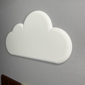Op de afbeelding: Een witte, wolk-vormige wanddecoratie met een afgeronde, getextureerde rand. De wolk is gemonteerd op een grijze muur, met een baksteenachtige structuur eronder zichtbaar. De wolk heeft een eenvoudig, strak ontwerp.