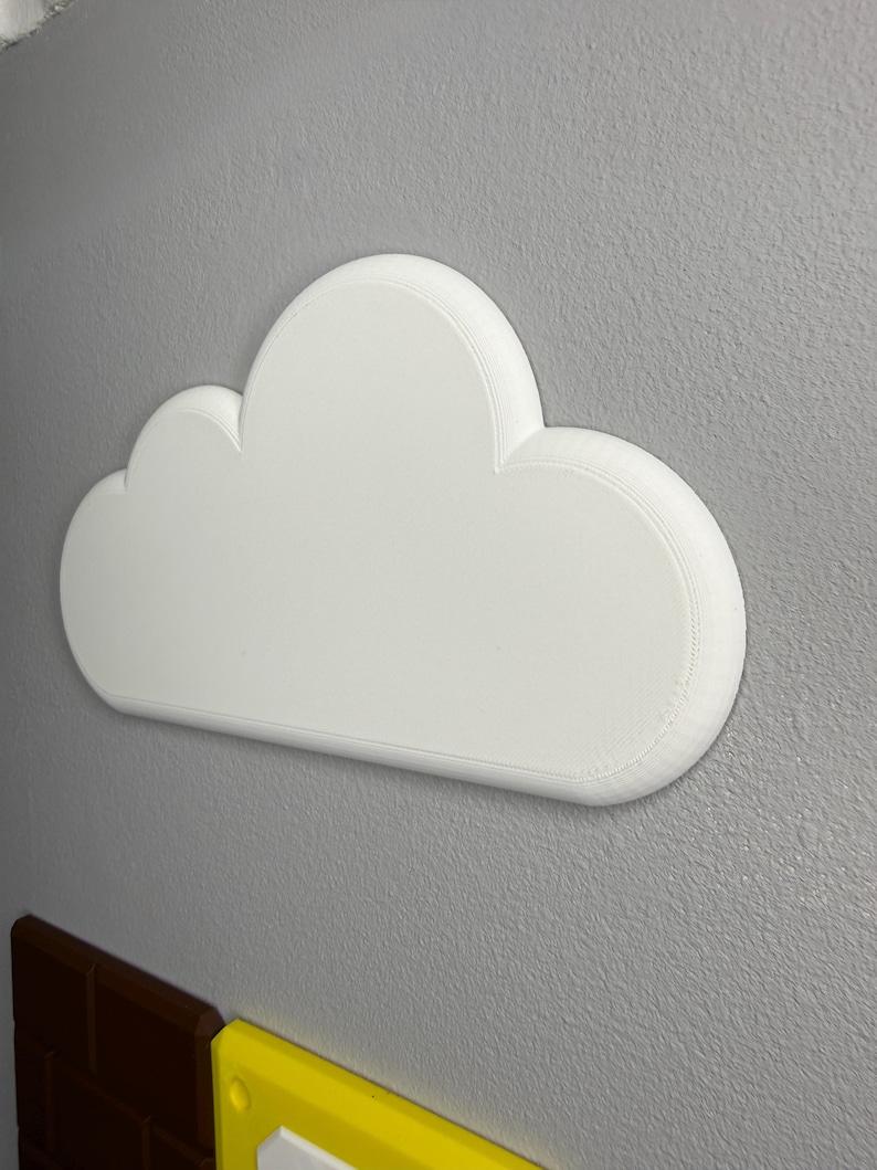 Op de afbeelding: Een witte, wolk-vormige wanddecoratie met een afgeronde, getextureerde rand. De wolk is gemonteerd op een grijze muur, met een baksteenachtige structuur eronder zichtbaar. De wolk heeft een eenvoudig, strak ontwerp.