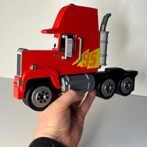 May include: A red toy semi-truck with the number 95 in yellow and black. The truck has a black chassis, silver exhaust pipe, and silver wheels. The truck is held in a person's hand.