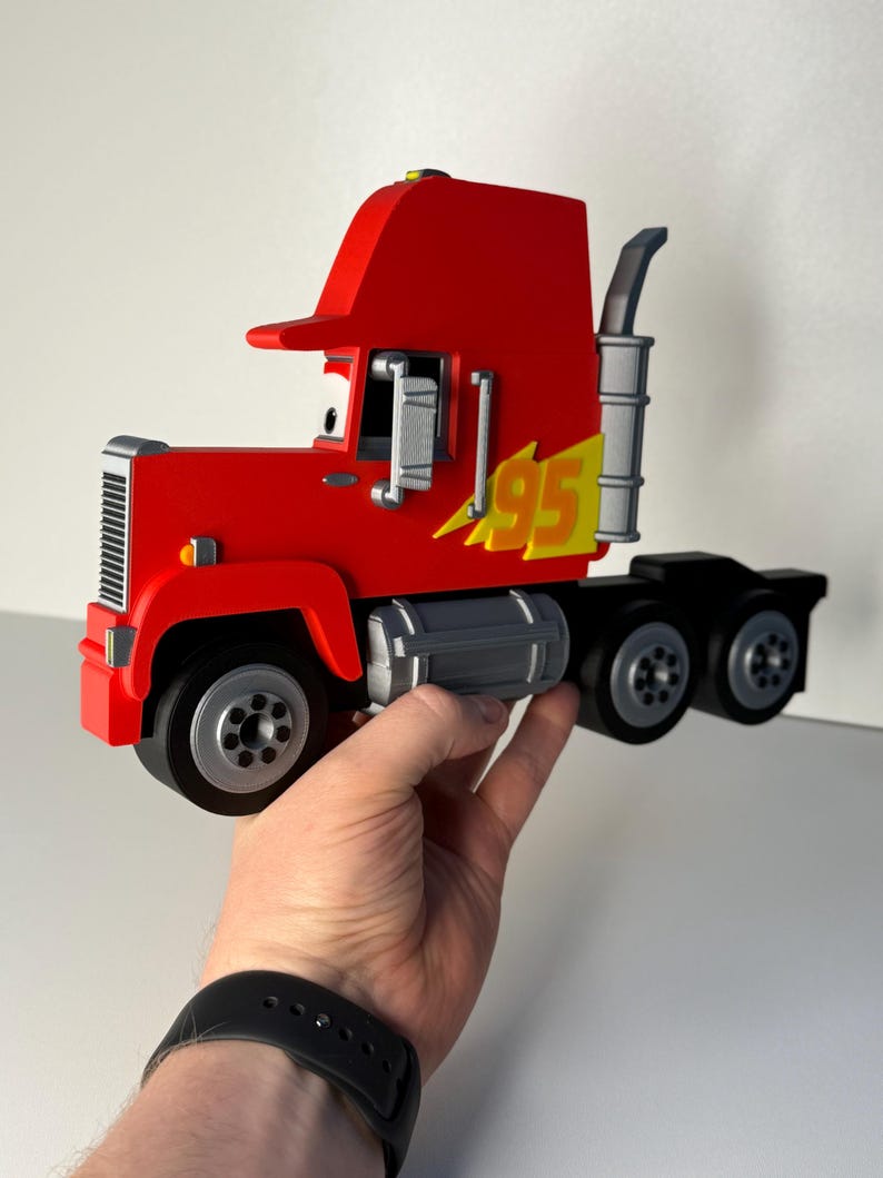 May include: A red toy semi-truck with the number 95 in yellow and black. The truck has a black chassis, silver exhaust pipe, and silver wheels. The truck is held in a person's hand.