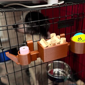 Brown PETG 3D-printed dog crate organiser for walkers and pet owners. Holds treats, tennis ball, and poo bags with slots for two leads. Heat-resistant, tool-free setup with adjustable hooks. Customisable with dog names, available in other colours.