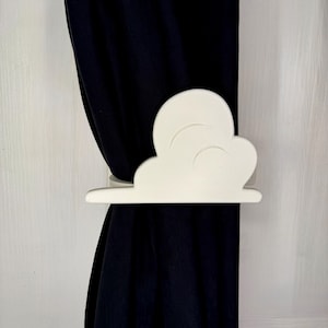 May include: A white cloud-shaped curtain tieback is wrapped around a dark curtain. The tieback is made of a solid, smooth material and has a three-dimensional design. The curtain is a solid, dark color. The background is a light color.
