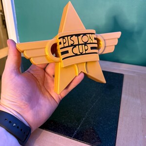 Puede incluir: Trofeo en forma de estrella, impreso en 3D, de color amarillo dorado. El trofeo presenta el texto "Piston Cup" en letras negras sobre una banda negra. El dise&ntilde;o incluye elementos en forma de alas y un pico triangular central.