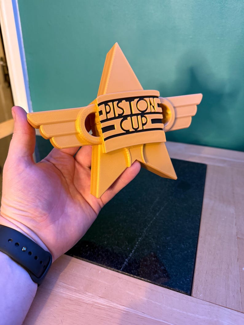 Puede incluir: Trofeo en forma de estrella, impreso en 3D, de color amarillo dorado. El trofeo presenta el texto "Piston Cup" en letras negras sobre una banda negra. El dise&ntilde;o incluye elementos en forma de alas y un pico triangular central.