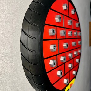 May include: A unique, wall-mounted motorcycle tire-shaped advent calendar. The black tire surrounds a red interior with silver-colored bolt-shaped compartments, each numbered. A yellow and black logo is visible at the bottom.