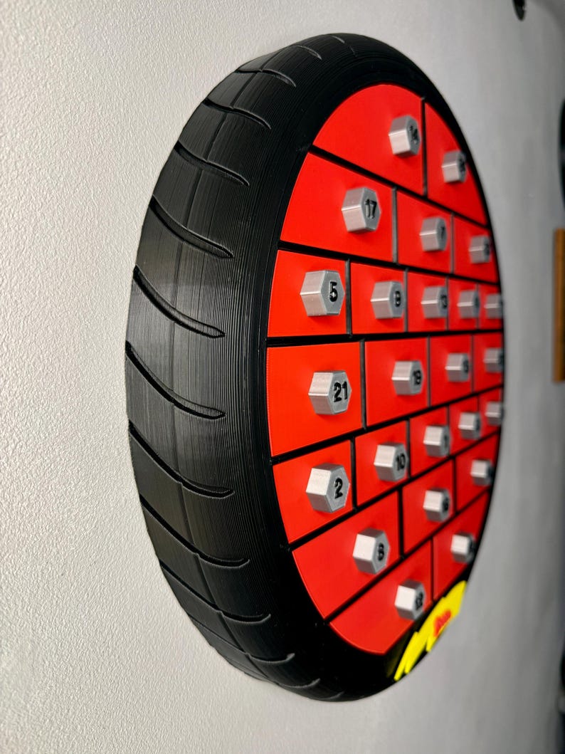 May include: A unique, wall-mounted motorcycle tire-shaped advent calendar. The black tire surrounds a red interior with silver-colored bolt-shaped compartments, each numbered. A yellow and black logo is visible at the bottom.