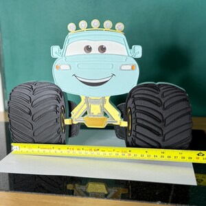 Op de afbeelding: Een lichtblauw monstertruckspeelgoed met grote zwarte banden en een lachend gezicht. De truck heeft gele en gouden accenten en een rij lampjes bovenop. Het speelgoed is ongeveer 30 cm lang.