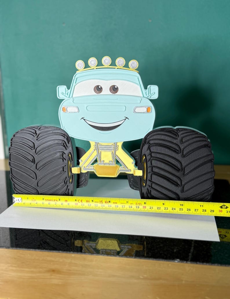 Op de afbeelding: Een lichtblauw monstertruckspeelgoed met grote zwarte banden en een lachend gezicht. De truck heeft gele en gouden accenten en een rij lampjes bovenop. Het speelgoed is ongeveer 30 cm lang.