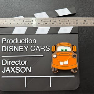 May include: A black clapperboard with white stripes and text that reads "Production DISNEY CARS Director JAXSON." An orange cartoon car is on the clapperboard. A ruler measures approximately 11 inches across the top.