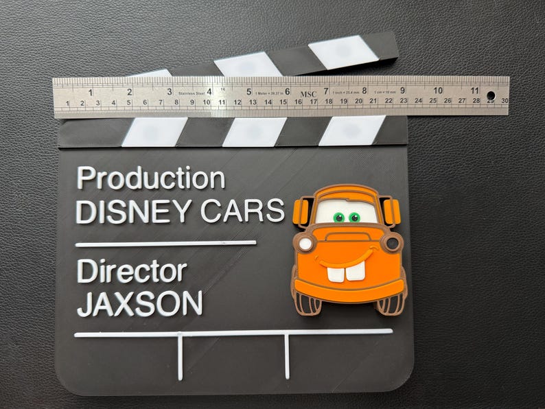 May include: A black clapperboard with white stripes and text that reads "Production DISNEY CARS Director JAXSON." An orange cartoon car is on the clapperboard. A ruler measures approximately 11 inches across the top.