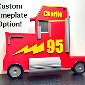 A 3D-printed red truck display inspired by Mack from Cars, featuring a bold yellow “95” and a lightning bolt. Silver details add realism, and a custom nameplate option is shown, reading “Charlie.”