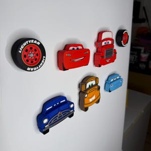 May include: A collection of colorful car-shaped magnets, including a red race car, a red truck, and a blue vintage car. A black tire magnet with the word "LIGHTYEAR" is also present. These magnets are attached to a white refrigerator.