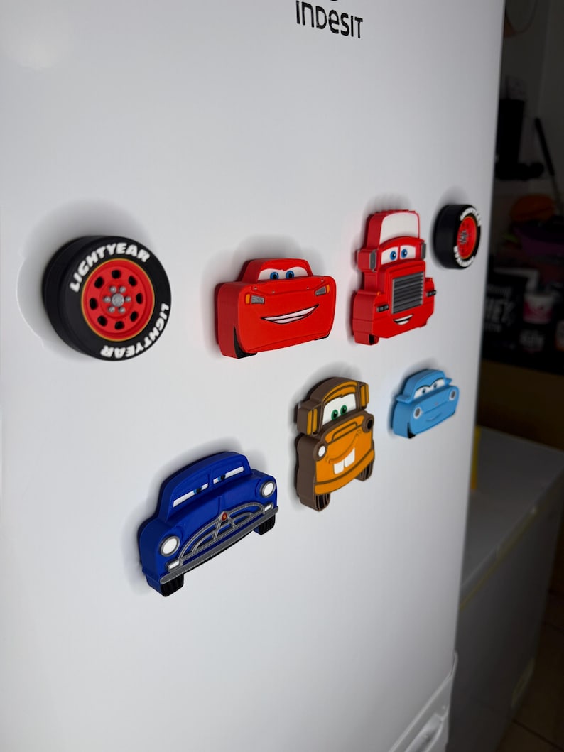 May include: A collection of colorful car-shaped magnets, including a red race car, a red truck, and a blue vintage car. A black tire magnet with the word "LIGHTYEAR" is also present. These magnets are attached to a white refrigerator.