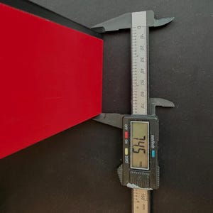 May include: A digital caliper measuring the thickness of a red object. The caliper displays a measurement of 7.45 mm. The tool is black with a digital display and inch/mm markings.