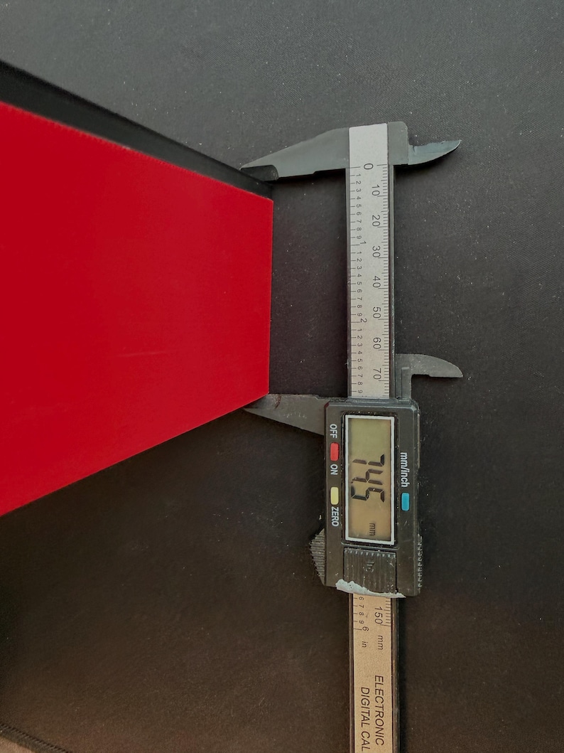 May include: A digital caliper measuring the thickness of a red object. The caliper displays a measurement of 7.45 mm. The tool is black with a digital display and inch/mm markings.