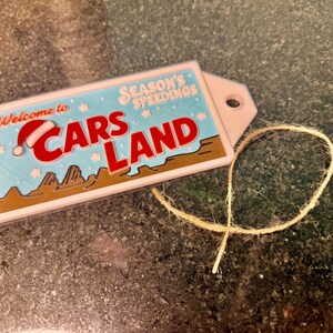 K&ouml;nnte beinhalten: Ein rechteckiges Etikett mit den Worten "Welcome to Cars Land" in Rot und Wei&szlig;, mit einem Weihnachtsmannhut auf dem Buchstaben "C". Das Etikett hat einen hellblauen Hintergrund mit den Worten "Season's Speedings". Ein beigefarbener Faden ist befestigt.