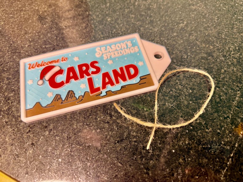 K&ouml;nnte beinhalten: Ein rechteckiges Etikett mit den Worten "Welcome to Cars Land" in Rot und Wei&szlig;, mit einem Weihnachtsmannhut auf dem Buchstaben "C". Das Etikett hat einen hellblauen Hintergrund mit den Worten "Season's Speedings". Ein beigefarbener Faden ist befestigt.