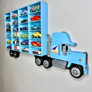 May include: A light blue wooden toy truck with a shelf for toy cars. The shelf is divided into multiple compartments, each holding a miniature car. The truck has black wheels and the word "Dinoco" on the side.