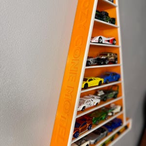 May include: A wall-mounted toy car display shelf in orange and white. The shelf is a tall, narrow rectangle with multiple levels. The text "COZY CONE MOTEL" is printed on the side. The shelf is filled with various toy cars. The shelf is mounted on a white wall.