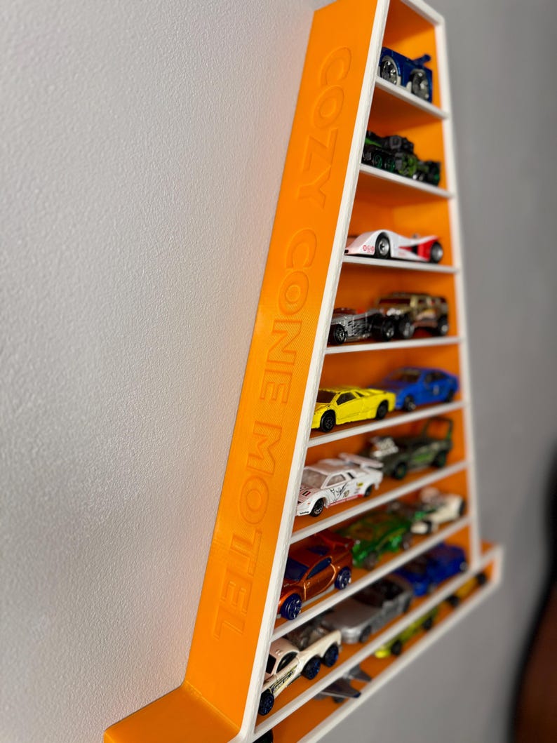 May include: A wall-mounted toy car display shelf in orange and white. The shelf is a tall, narrow rectangle with multiple levels. The text "COZY CONE MOTEL" is printed on the side. The shelf is filled with various toy cars. The shelf is mounted on a white wall.