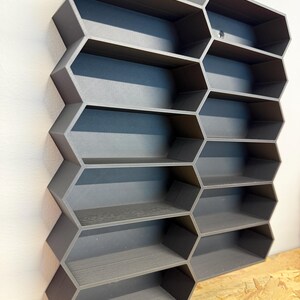 May include: A dark gray wall-mounted storage unit featuring a modular honeycomb design with multiple hexagonal compartments. The structure is composed of interconnected geometric cells arranged in a vertical grid, providing open shelving space for small items.