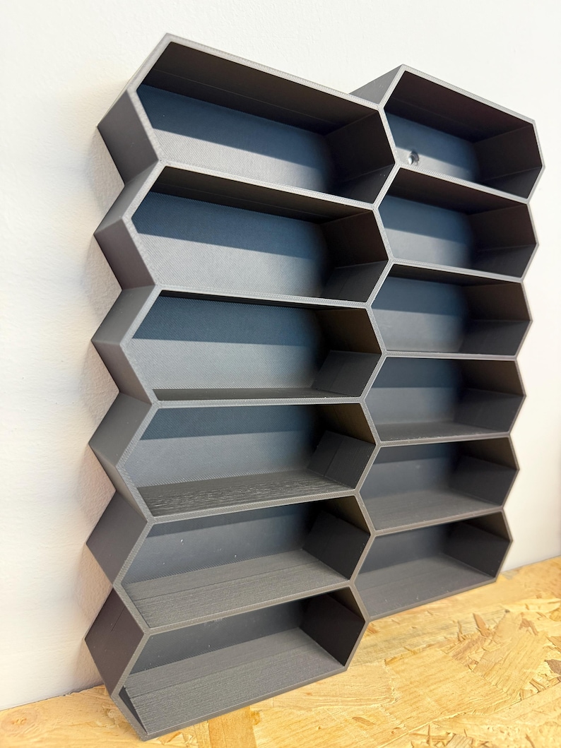 May include: A dark gray wall-mounted storage unit featuring a modular honeycomb design with multiple hexagonal compartments. The structure is composed of interconnected geometric cells arranged in a vertical grid, providing open shelving space for small items.