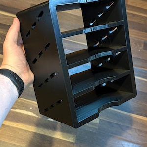 May include: A black, multi-tiered organizer with four shelves. The organizer has a rectangular shape with angled cutouts on the sides. The item is held by a person's hand.