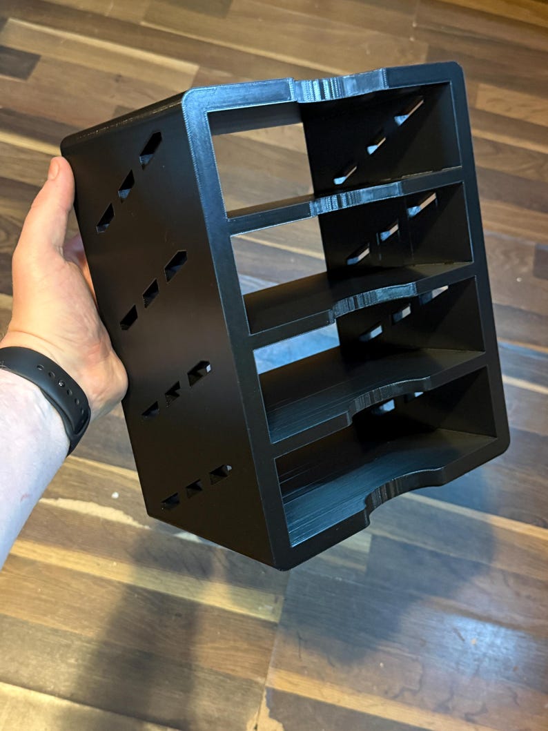 May include: A black, multi-tiered organizer with four shelves. The organizer has a rectangular shape with angled cutouts on the sides. The item is held by a person's hand.