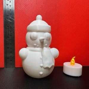 White 3D-printed snowman LED tealight holder displayed against a red background with a ruler showing dimensions and an LED tealight placed nearby for size reference.