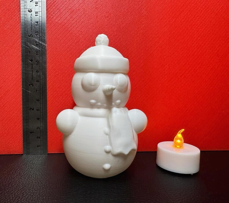White 3D-printed snowman LED tealight holder displayed against a red background with a ruler showing dimensions and an LED tealight placed nearby for size reference.