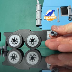 May include: A light blue toy truck cab with a Dinoco logo and a black trailer section with wheels. A hand holds a small gray cylinder, possibly a part of the truck. The toy is on a reflective surface.