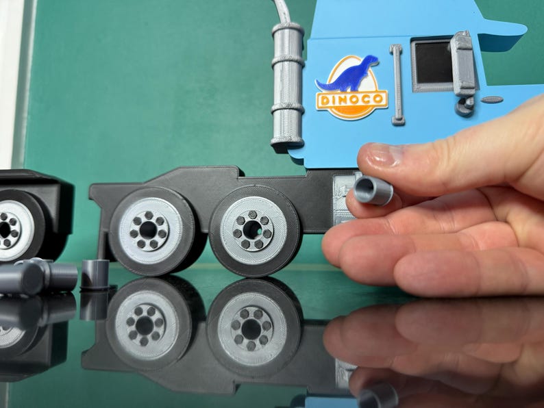 May include: A light blue toy truck cab with a Dinoco logo and a black trailer section with wheels. A hand holds a small gray cylinder, possibly a part of the truck. The toy is on a reflective surface.