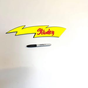 May include: A yellow arrow-shaped sign with the name "Stanley" in red script. A black and silver Sharpie marker is below the sign. The sign has a black outline and a lightning bolt design.