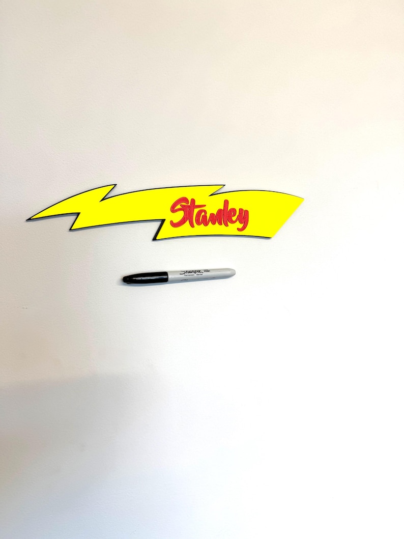 May include: A yellow arrow-shaped sign with the name "Stanley" in red script. A black and silver Sharpie marker is below the sign. The sign has a black outline and a lightning bolt design.