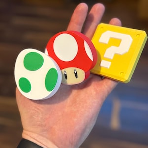 May include: A collection of colorful 3D-printed items from the Super Mario video game series. Includes a red mushroom with a white top, a green and white egg, and a yellow question mark block. The items are held in a hand.