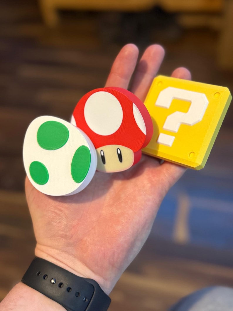May include: A collection of colorful 3D-printed items from the Super Mario video game series. Includes a red mushroom with a white top, a green and white egg, and a yellow question mark block. The items are held in a hand.